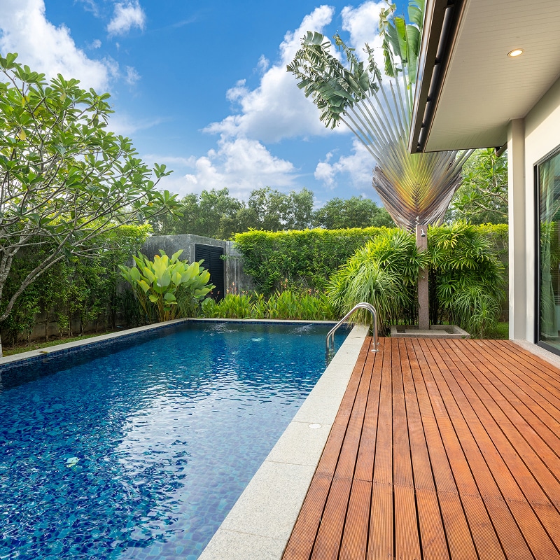 Piscine de villa moderne avec terrasse en bois entourée d'un jardin tropical verdoyant sous un ciel bleu.